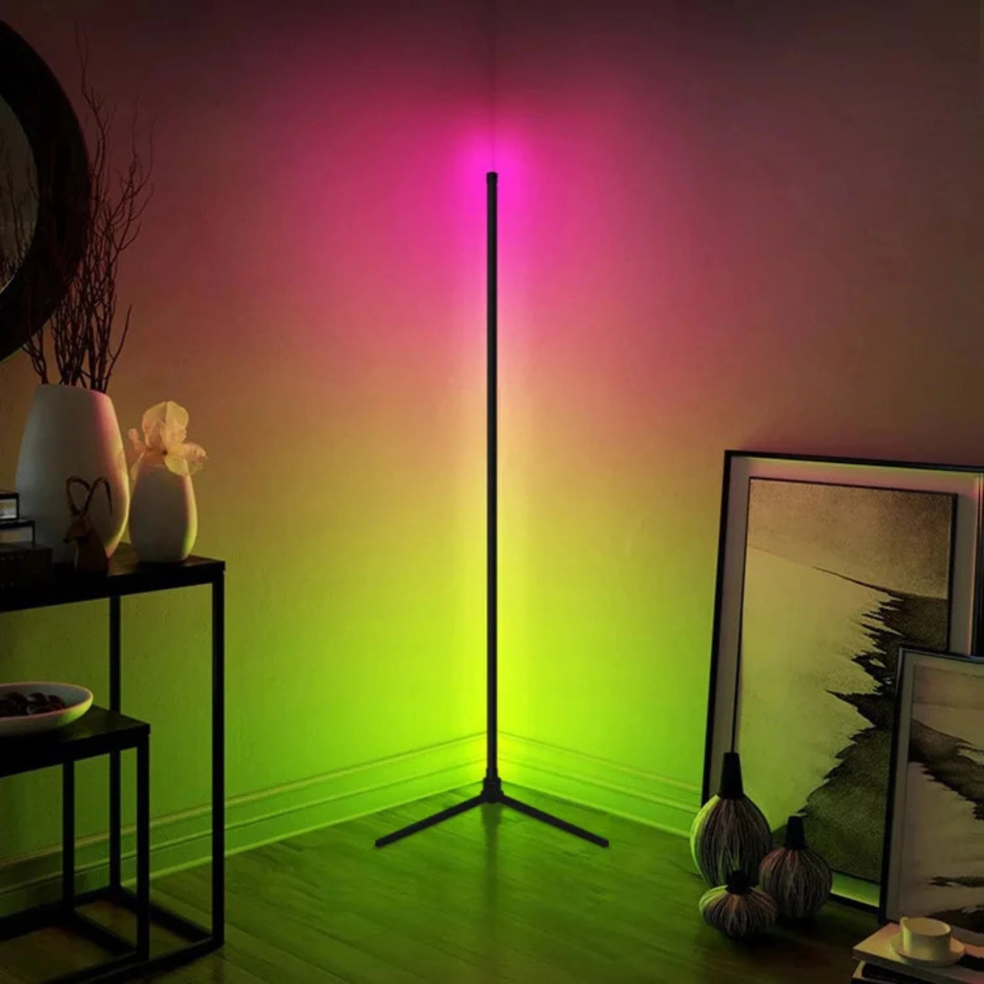 A modern corner floor lamp emitting vibrant pink and green light, creating a colorful and ambient glow in a living room. The lamp has a sleek, black design with a thin stand and minimalist structure. It is positioned in the corner of the room, illuminating the surrounding walls and floor. The room features a small black table with decorative items, a mirror on the wall, and framed artwork leaning against the wall. The overall setting is cozy and stylish, showcasing the lamp's ability to enhance the atmosphe